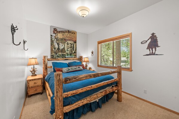 Une chambre à coucher avec un lit en bois, une affiche accrochée au mur, une fenêtre avec des stores et une table de chevet avec une lampe.