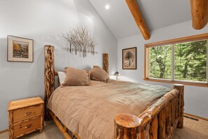 Een slaapkamer met een houten bedframe, een nachtkastje, een lamp en een raam met uitzicht op bomen.