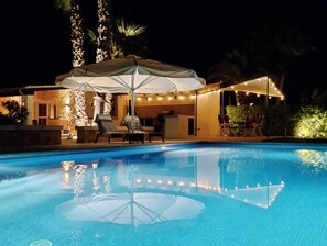 Une piscine entourée d’un mur de pierre et de palmiers, avec des chaises longues et un parasol.