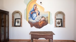 A mural with a religious theme, a wooden bench, and two arched niches with framed pictures. - La Casa Del Priore (Patti)