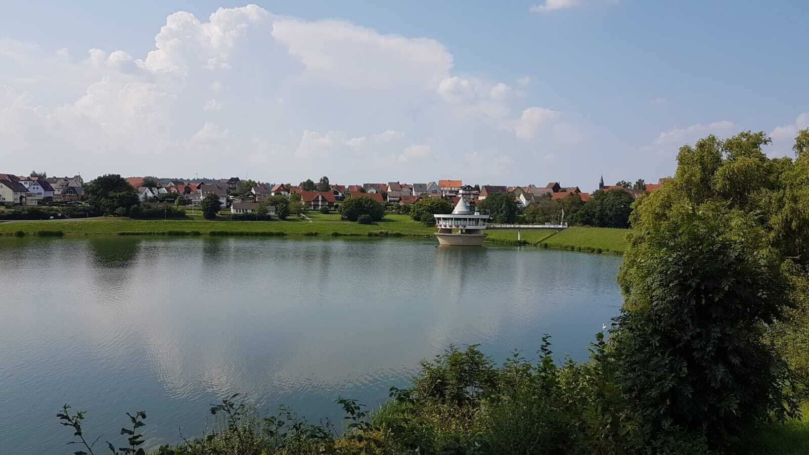 A calm lake with a small boat in the center, surrounded by greenery and a town in the background.