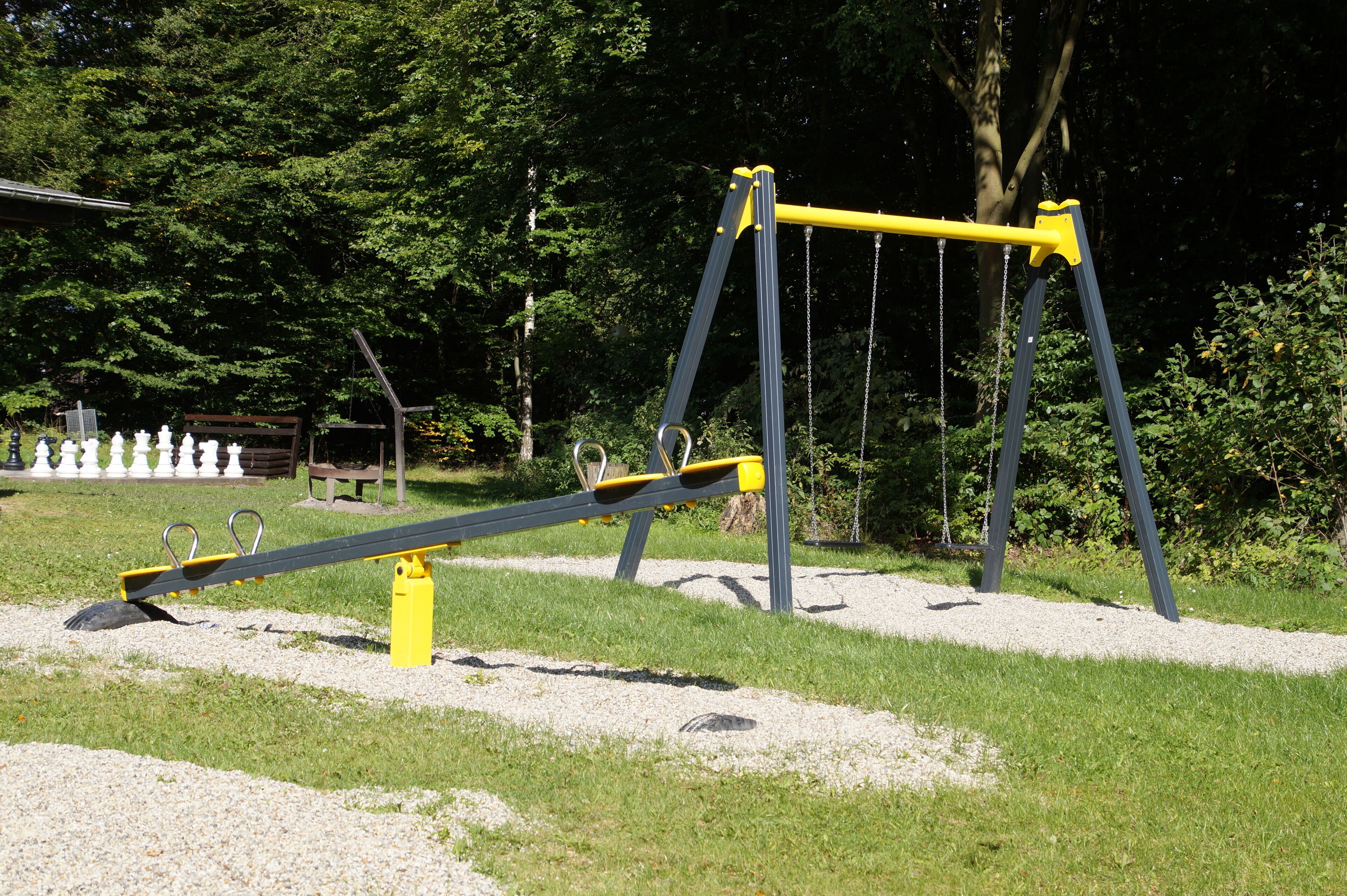 Un sube y baja de colores amarillo y gris en un parque con árboles y una valla blanca al fondo.