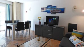 Una sala moderna con una mesa de centro de vidrio, un televisor en un mueble bajo, un comedor con sillas y un sofá con un cojín colorido.