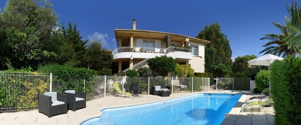 Uma casa de dois andares com piscina, cercada por um muro branco e diversos móveis de jardim.