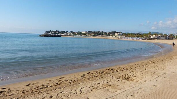 A beach with clear blue water, a sandy shore, and a distant shoreline with buildings and trees.