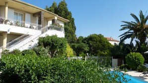A two-story house with a balcony, surrounded by lush greenery and a swimming pool.