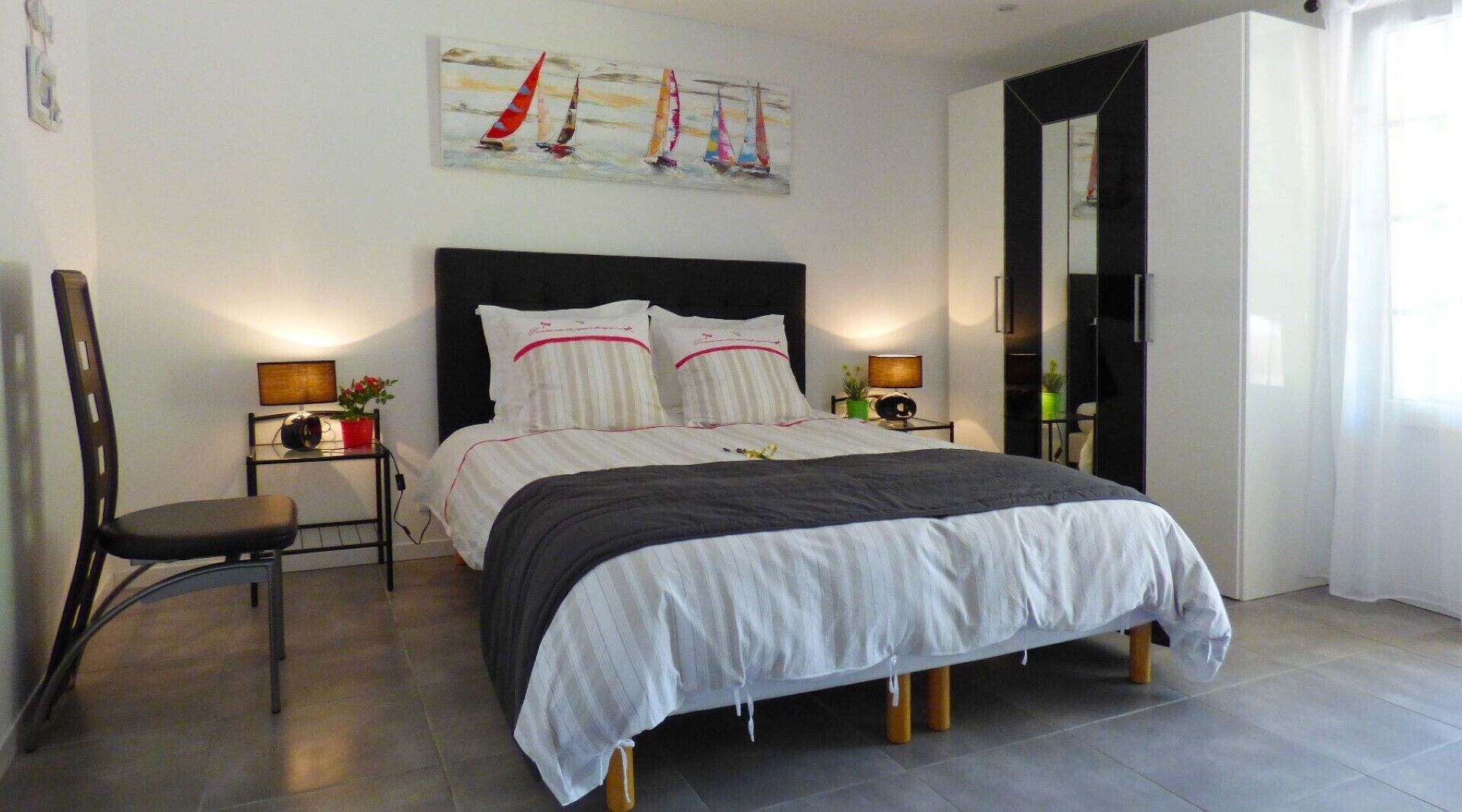 Un dormitorio moderno con una cama grande, dos mesitas de noche con lámparas, una silla y un cuadro con barcos a vela en la pared.