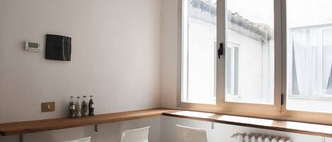 A corner room with a wooden shelf, two white chairs, a radiator, and a window with a view of a building outside.