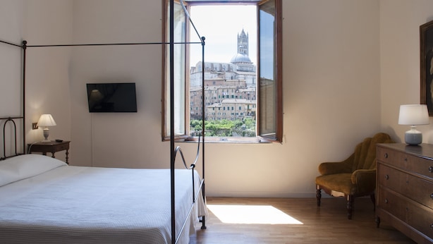 A bedroom with a four-poster bed, a wooden dresser, a chair, and a window with a cityscape view. - La Sapienza Di Al (Siena)