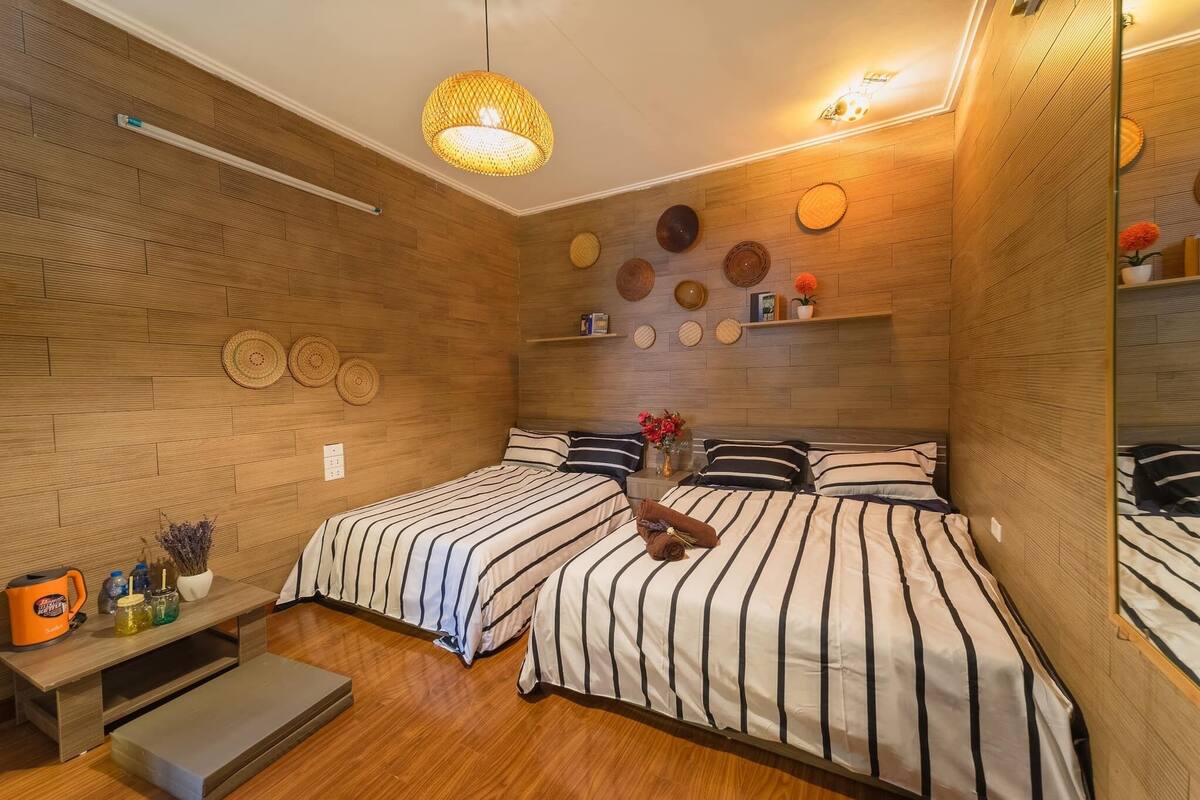 A hotel room with two beds, wooden flooring, a hanging light fixture, and wall-mounted shelves with decorative items.