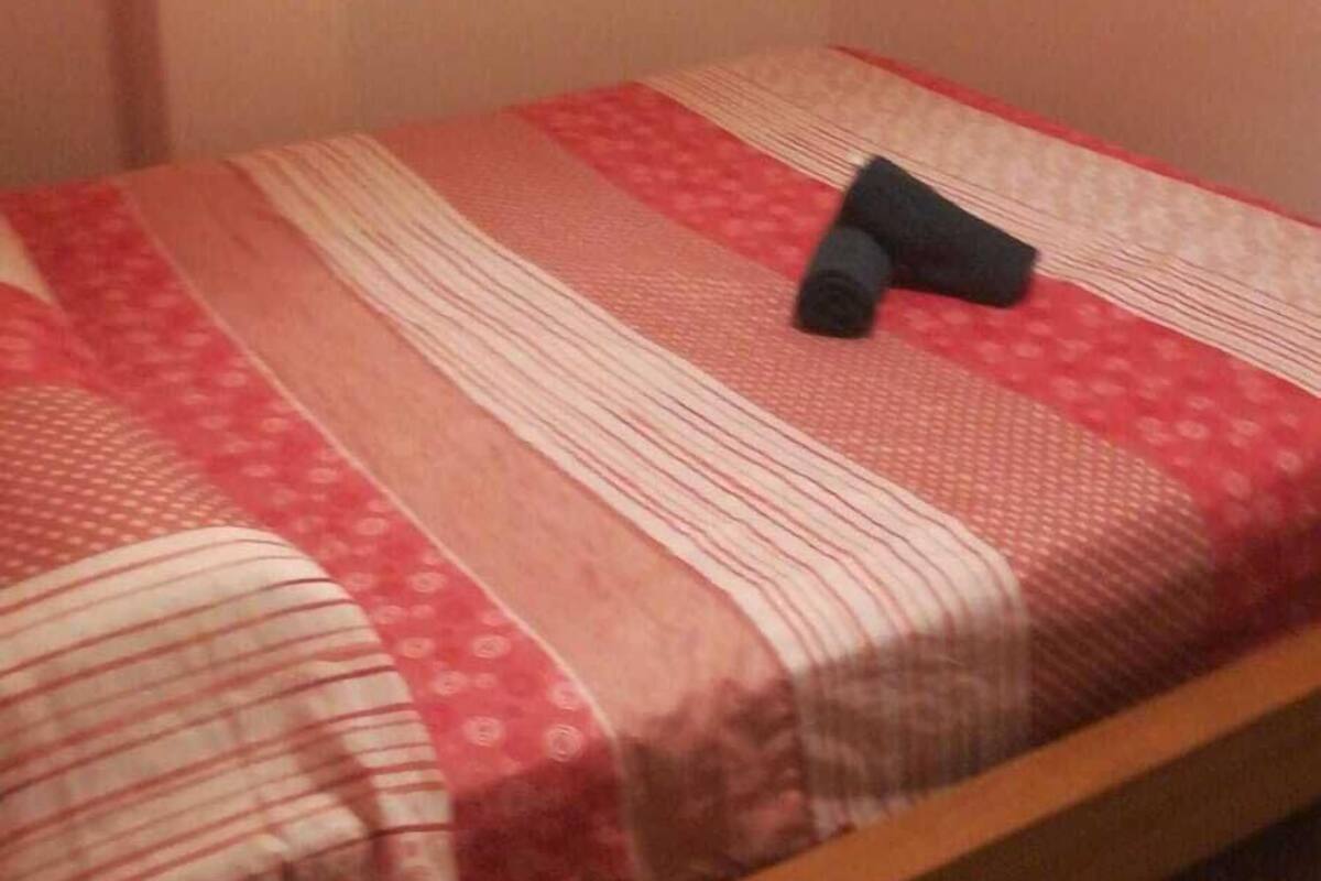 Una habitación pequeña con cama individual, estructura de madera, colcha a rayas roja y blanca, y una manta oscura enrollada sobre la cama.