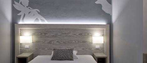 A modern bedroom with a bed, two bedside tables, a headboard with integrated lighting, and wall art depicting a fairy and a deer.