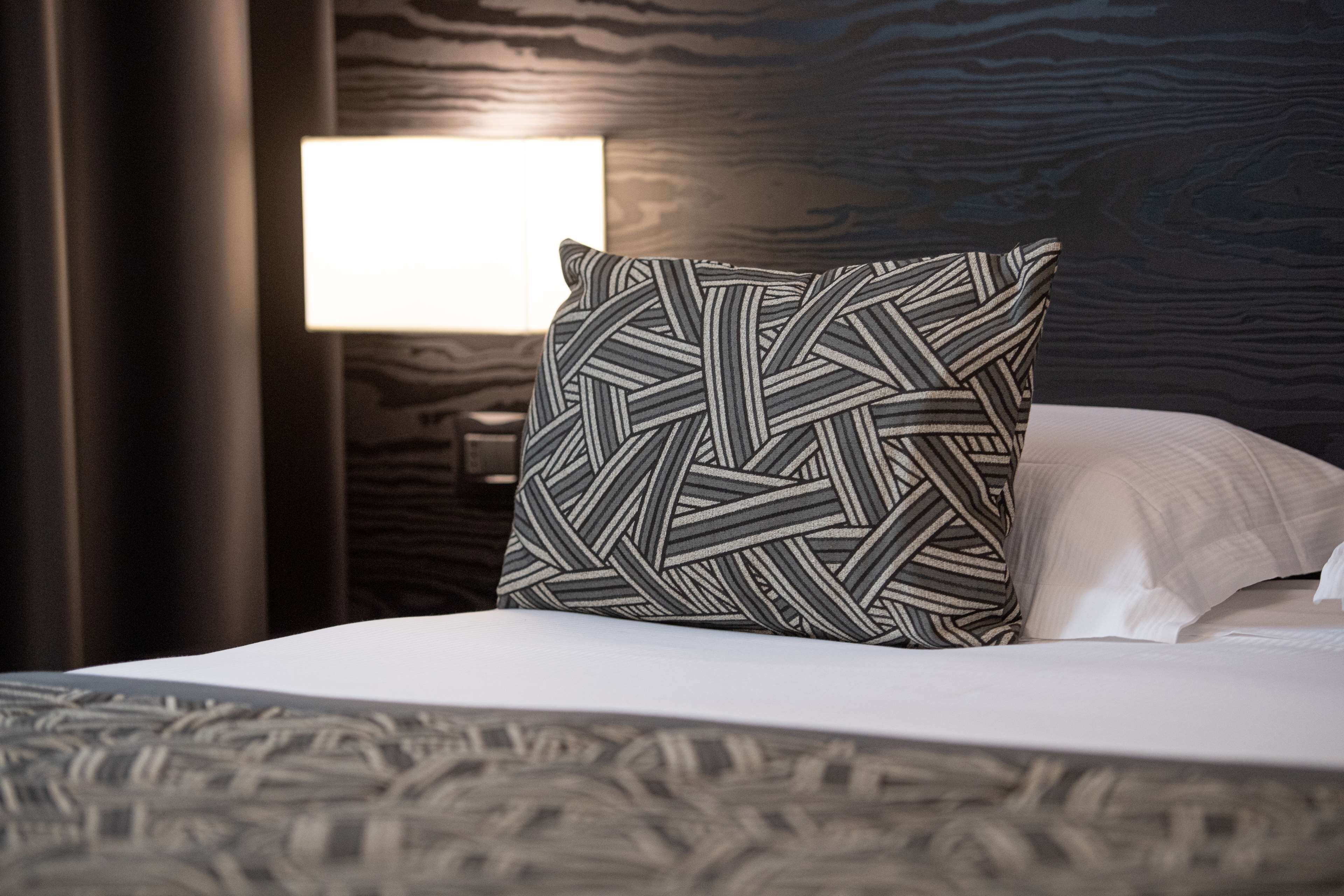 Una cama bien tendida con un almohadón estampado y uno liso, una lámpara de noche y un cabecero de madera oscura.