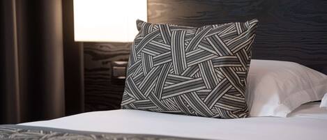 Una cama bien tendida con un almohadón estampado y uno liso, una lámpara de noche y un cabecero de madera oscura.
