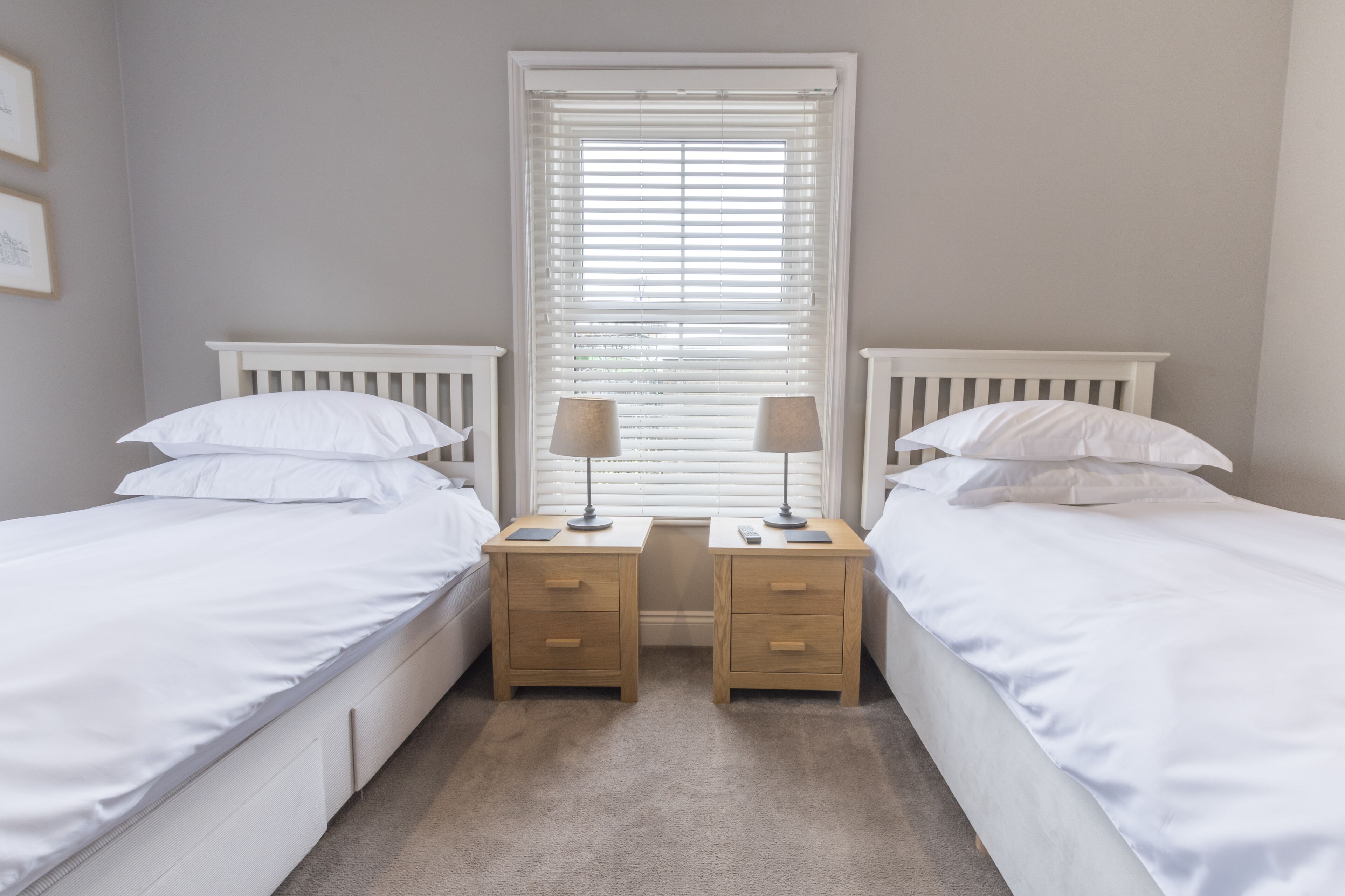 A twin bedroom with two single beds, a window with blinds, bedside tables with lamps, and a carpeted floor.