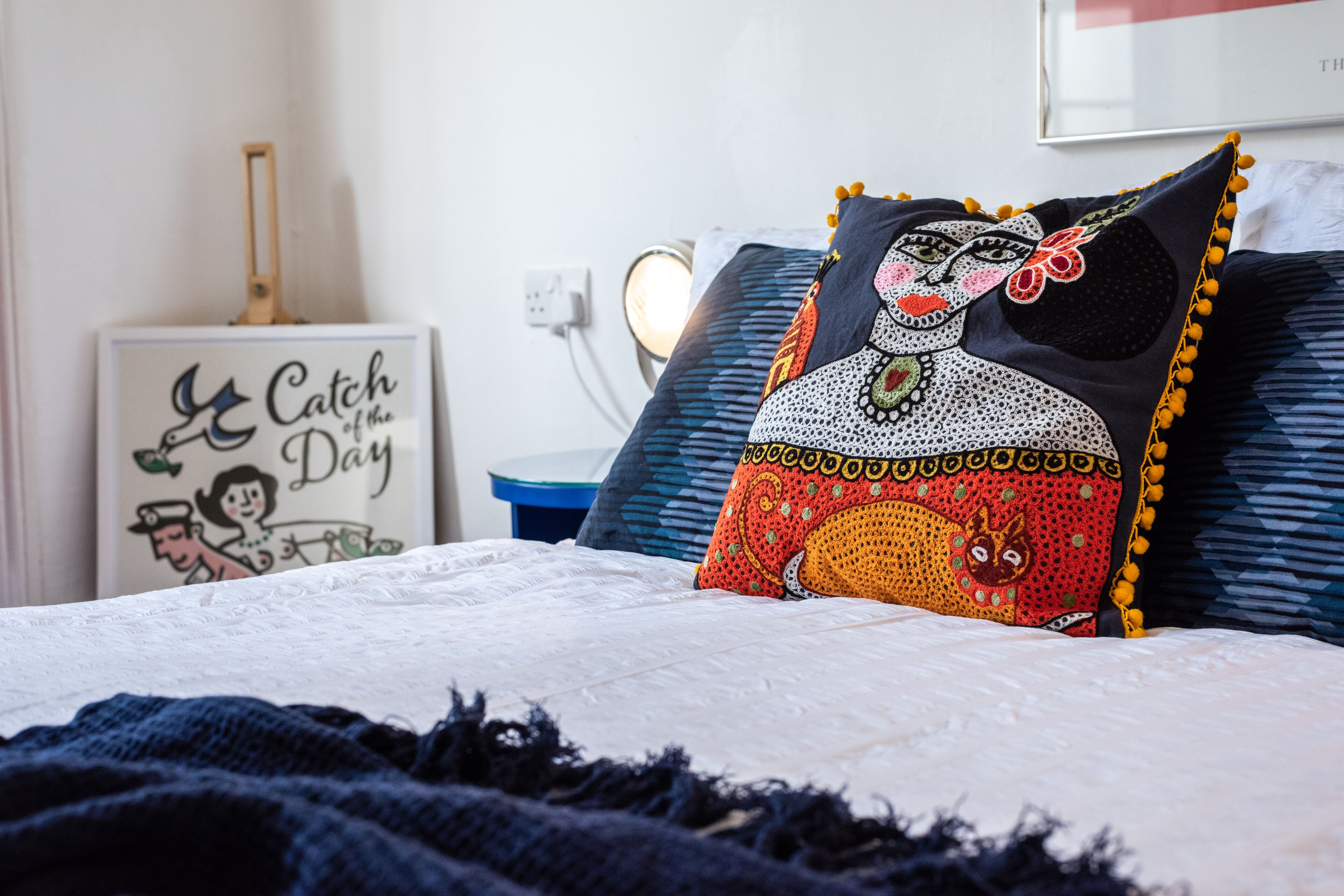 Ein Bett mit weißer Decke, ein dekoratives Kissen mit farbigem Muster und ein Nachttisch mit Lampe.