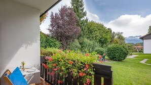 Un balcón con una silla y mesa de madera, un jardín con valla de madera y vistas a una cadena montañosa.
