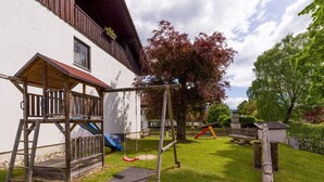 Estructura de juego de madera con tobogán y columpio, una pequeña terraza de madera, y un patio cercado con árboles y un edificio al fondo.