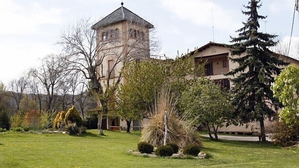 Ein Gebäude mit einem Turm, umgeben von einem gepflegten Garten mit verschiedenen Pflanzen und Bäumen.