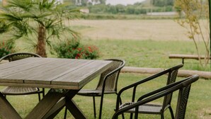 Eine Terrasse im Freien mit einem Holztisch und Korbstühlen, umgeben von Grünpflanzen und Blick auf ein Feld und ferne Hügel.