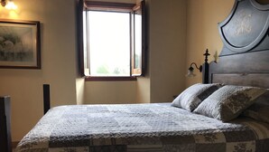 Ein ordentlich bezogenes Bett mit gesteppter Decke, ein Fenster mit Blick auf Grün und eine Wandleuchte.