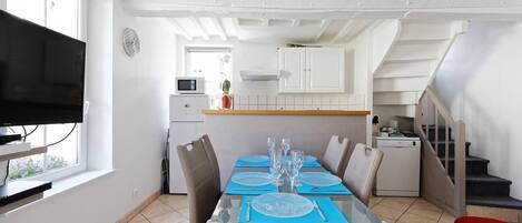 Un coin repas avec une table dressée pour quatre personnes, un téléviseur sur le côté gauche et une cuisine en arrière-plan.