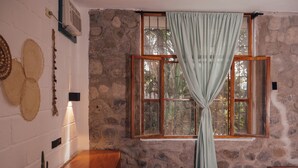 Una habitación con pared de piedra, una cama de madera, una mesa y un ventanal con cortinas.