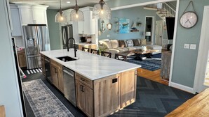 Uma cozinha moderna com uma ilha grande, eletrodomésticos de aço inoxidável, iluminação pendente e uma sala de estar aconchegante com sofá e mesa de centro.
