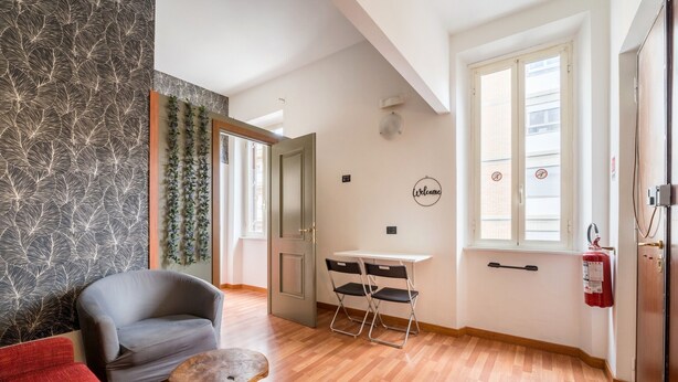 A hotel room with a grey armchair, a red sofa, a small wooden table, a white desk with two black chairs, a fire extinguisher, and a 'Welcome' sign on the wall. - Rooms Piramide (Rome)
