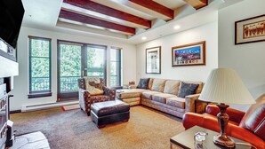A living room with a sectional sofa, a red leather chair, a coffee table with a lamp, and a fireplace. There are large windows with blinds, a fireplace, and framed pictures on the wall.