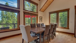 A dining area with a large wooden table and chairs, large windows with a view of trees, and a wooden ceiling.