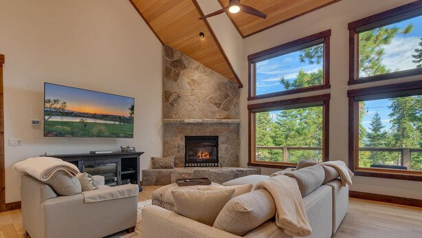 A living room with a stone fireplace, large windows, a flat-screen TV, and a cozy seating area.