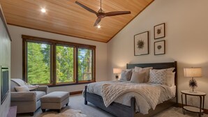 A bedroom with a large bed, a ceiling fan, a window with a view of trees, and a small table with a lamp.