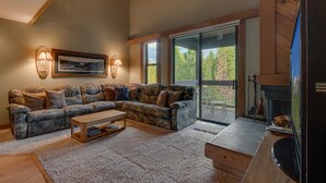A living room with a sectional sofa, a coffee table, and a fireplace. There is a large sliding glass door leading to a deck with outdoor furniture.