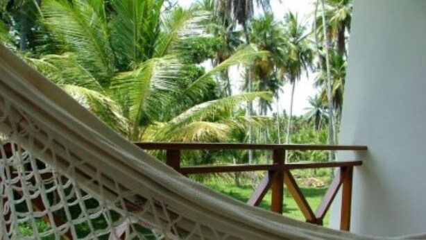 A hammock with a wooden railing, set against a backdrop of lush green palm trees. - Garapuá Praia Hotel (Cairu)