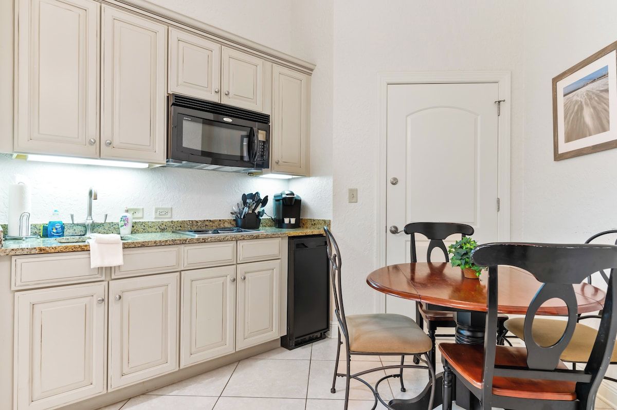 Une cuisine avec des meubles de couleur beige, un four à micro-ondes, une machine à café et un lave-vaisselle. On y trouve une table à manger avec des chaises et une plante en pot.