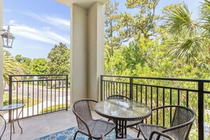 A balcony with a glass table and wicker chairs, overlooking a road and lush greenery. - Beach Air -optional BIKES-Pool-Walk 2 Pier Village (St Simons)