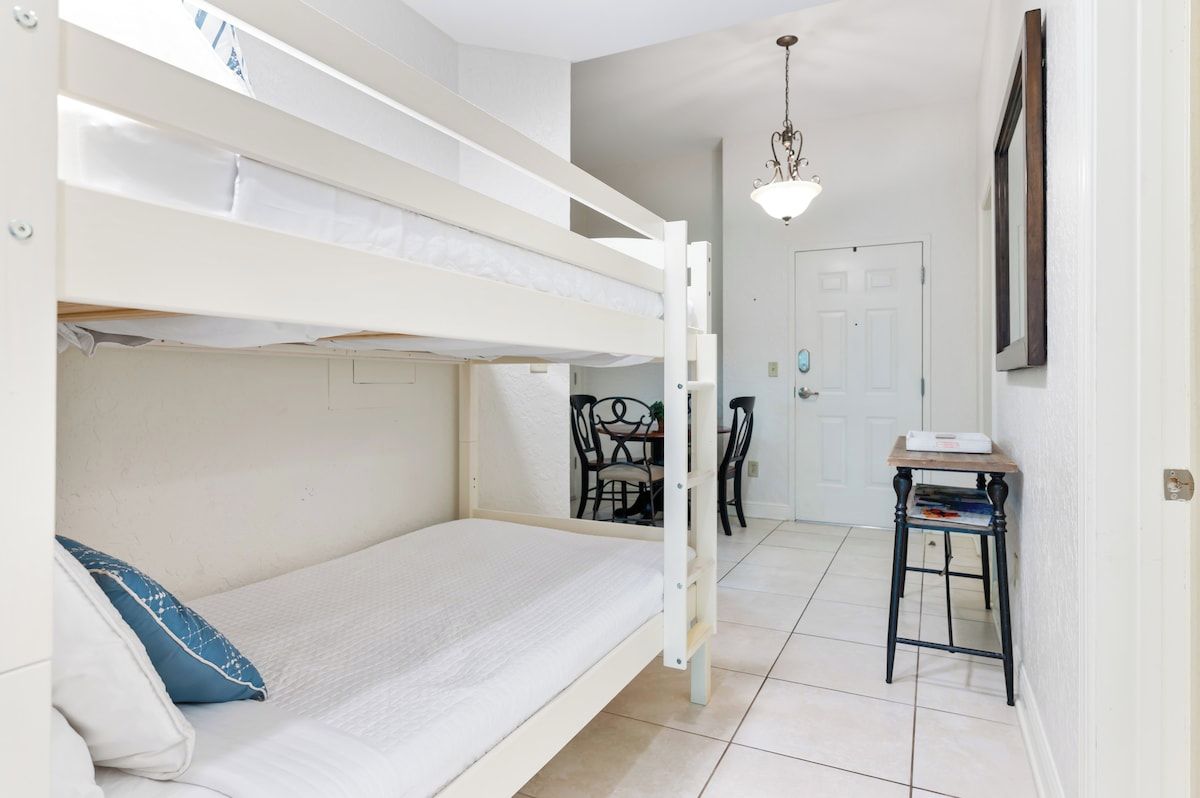 A bunk bed room with a desk and chairs, a door, and a hanging light fixture.