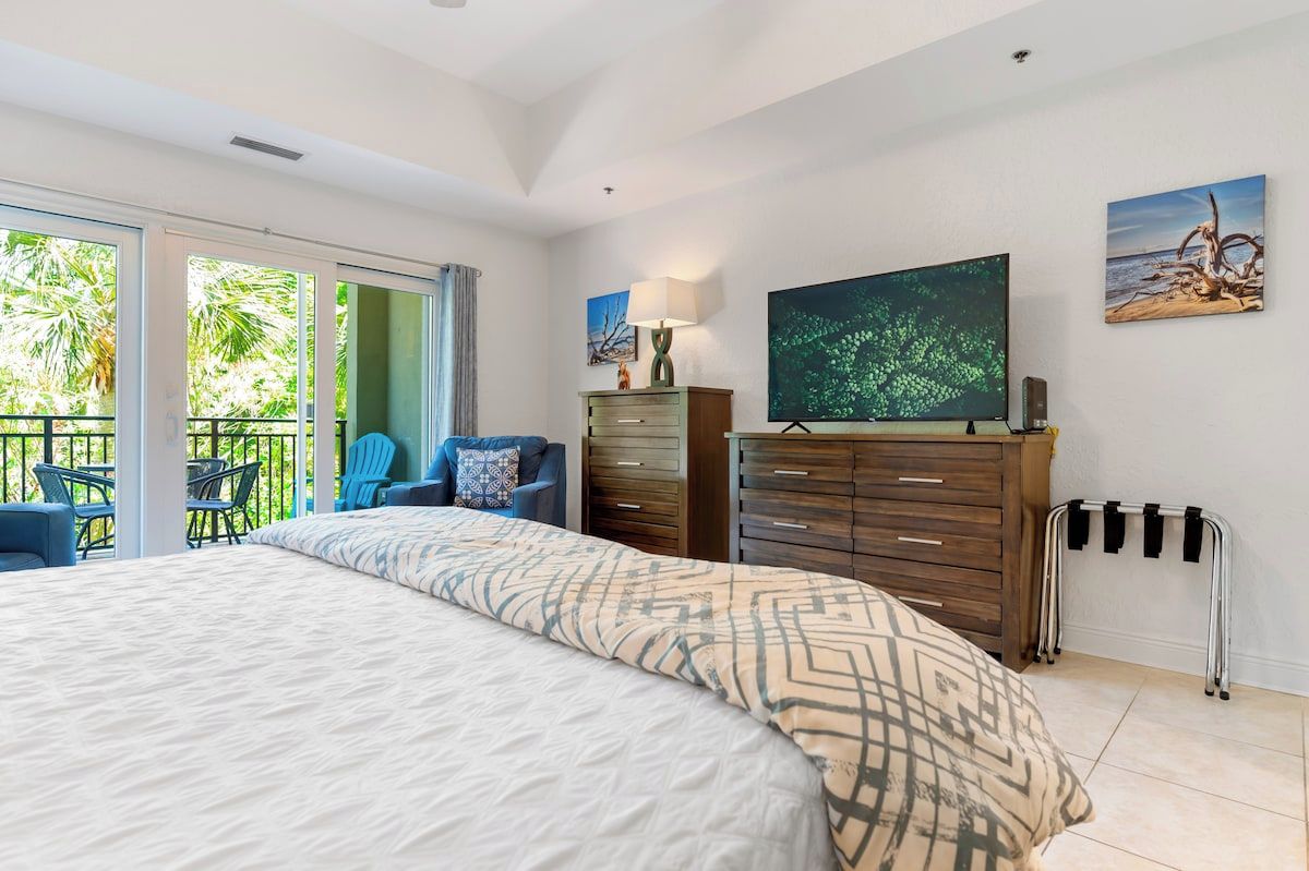 Une chambre avec un grand lit, une télévision, une commode et un balcon équipé de chaises.
