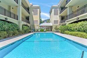 A swimming pool with a view of a building with balconies and a covered entrance. - Beach Air -optional BIKES-Pool-Walk 2 Pier Village (St Simons)