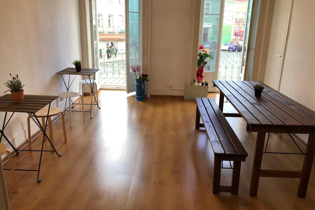 A room with wooden flooring, two tables, and benches, with a view of a balcony and buildings outside.