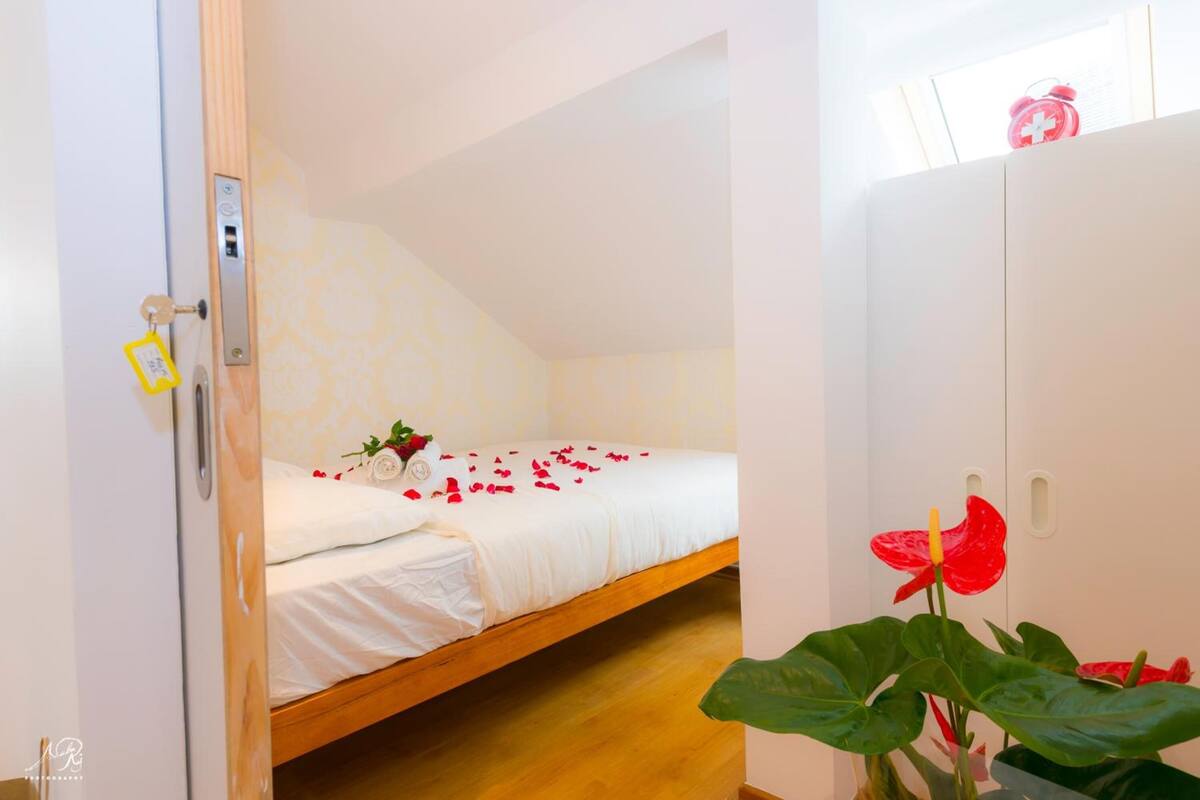 Una cama individual con ropa de cama blanca y pétalos de rosa, una puerta de madera con cerradura de llave y una flor roja en maceta.