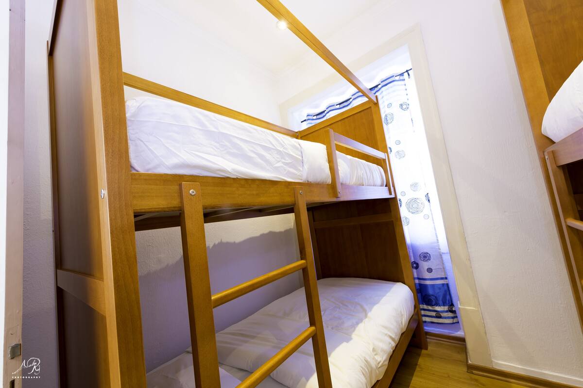 Habitación con literas de madera, ropa de cama blanca y una cortina con estampado azul y blanco.