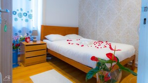 Ein ordentlich bezogenes Bett mit weißen Leinentüchern und Rosenblättern, ein Nachttisch aus Holz mit einer Lampe, eine Vase mit Blumen und eine Wand mit Muster.