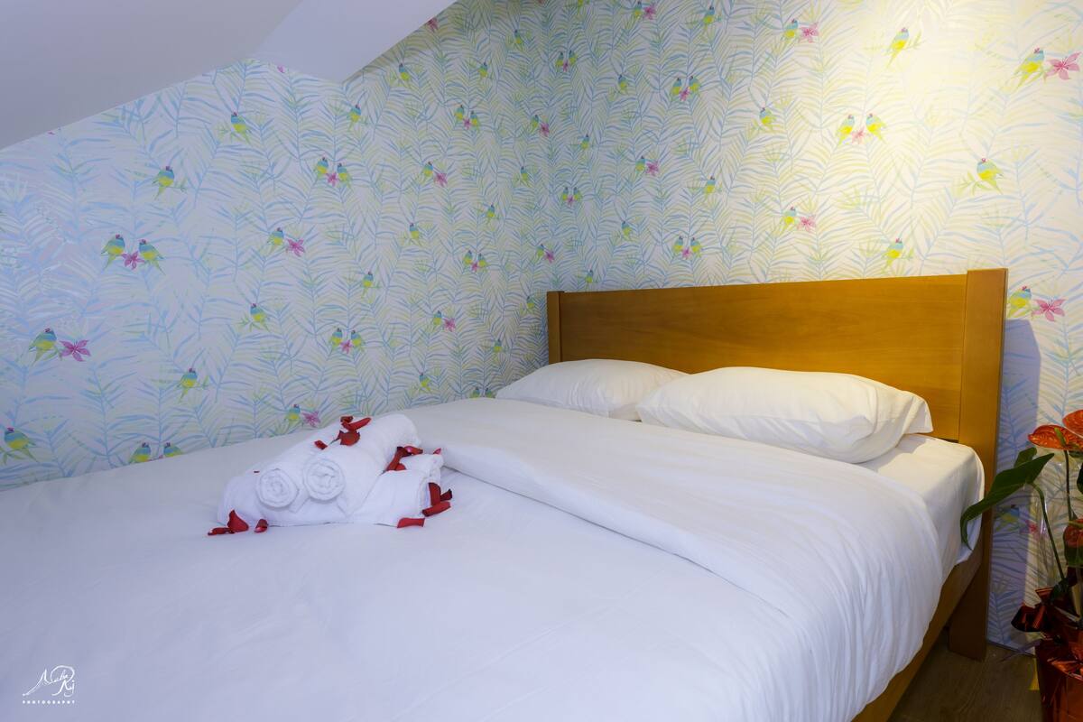 Una cama impecablemente hecha con ropa blanca y detalles rojos, un cabecero de madera y un estampado de flores en el papel tapiz.