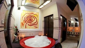 A modern bathroom with a large, circular, red bathtub filled with bubbles, a large abstract painting on the wall, and a mirror above the sink. - Hotel & Motel Le Baron (Porto Ferreira)