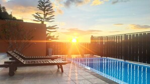Un espace piscine aménagé sur le toit, avec un fauteuil de relaxation en bois, un petit arbre et une vue sur le coucher de soleil sur l’eau.