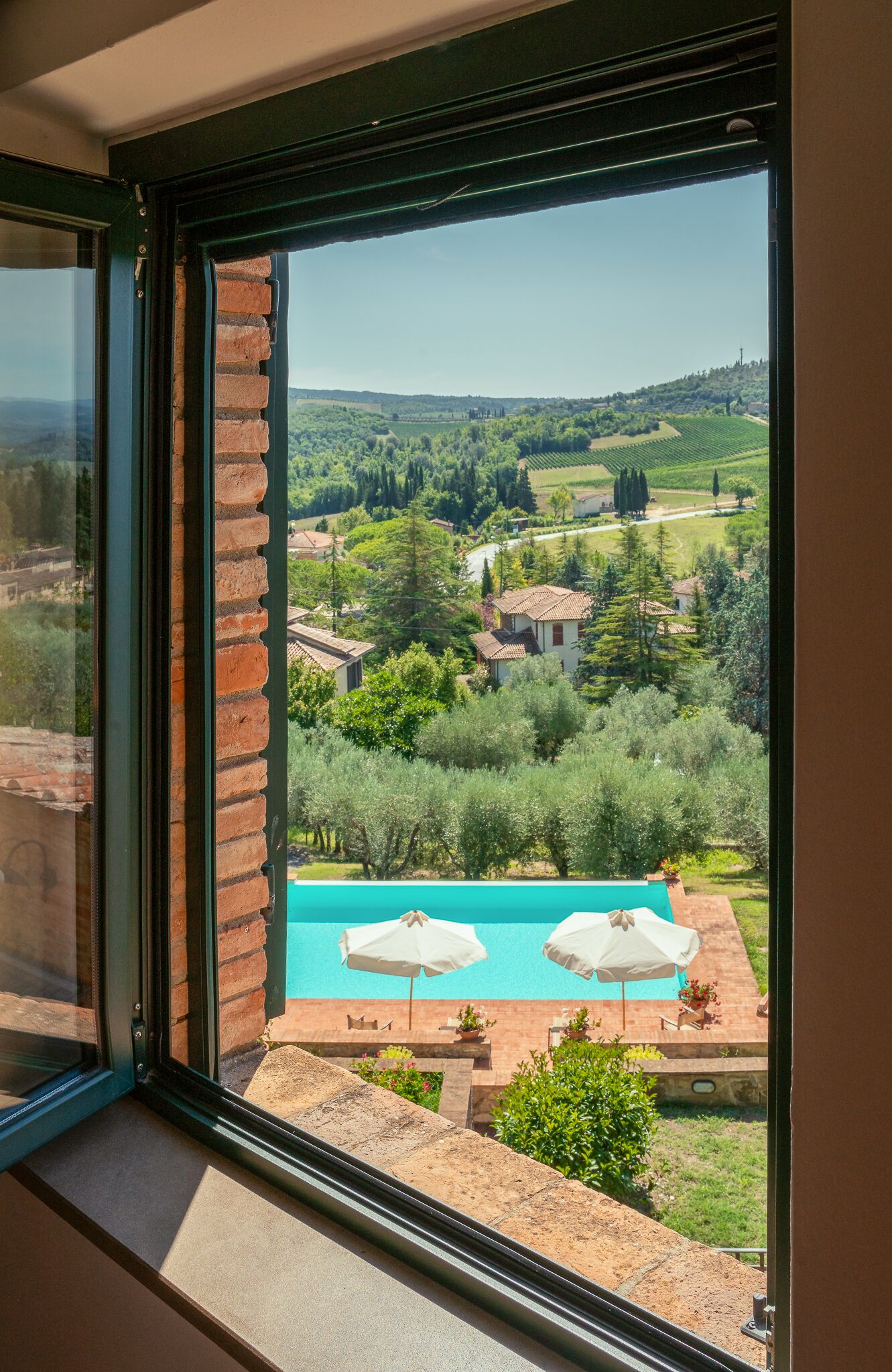 Blick aus einem Fenster auf einen Swimmingpool mit zwei Sonnenschirmen, umgeben von viel Grün und einer ländlichen Landschaft.