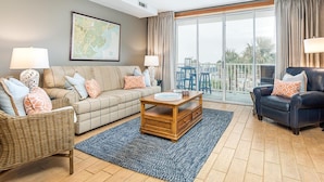 A living room with a sofa, armchairs, a coffee table, a map on the wall, and a view through large windows. - Tybee Beach Club 125 | Easy Walk to Beach + Pool! (Tybee Island)