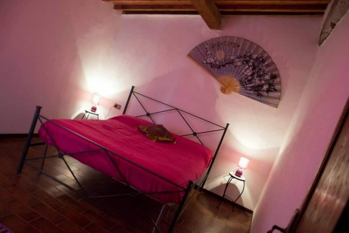 Ein Einzelbett mit Metallgestell und roter Bettdecke in einem Raum mit Holzbalkendecke. Ein an der Wand befestigter Ventilator mit floralem Dekor und zwei Nachttischlampen stehen auf dem Boden.